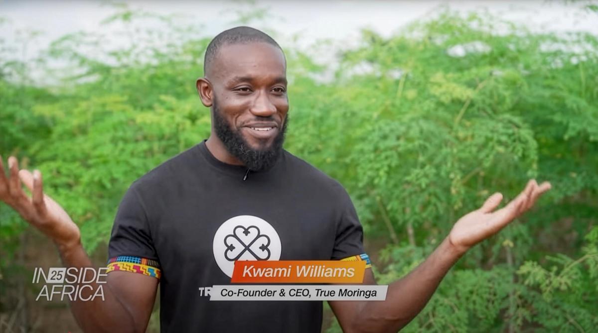 Kwami Williams, Co-Founder and CEO of True Moringa, speaks on CNN’s Inside Africa program. He smiles with arms raised slightly, standing outdoors in front of lush green moringa plants.